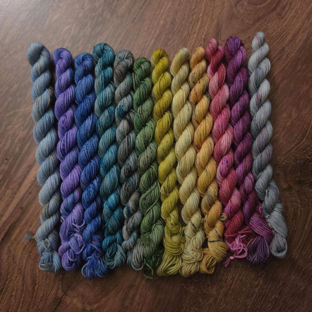 Twelve twisted yarn skeins arranged in a fan shape on a dark brown wooden surface, displaying a rainbow of colors from blue and purple to green, yellow, pink, and gray.