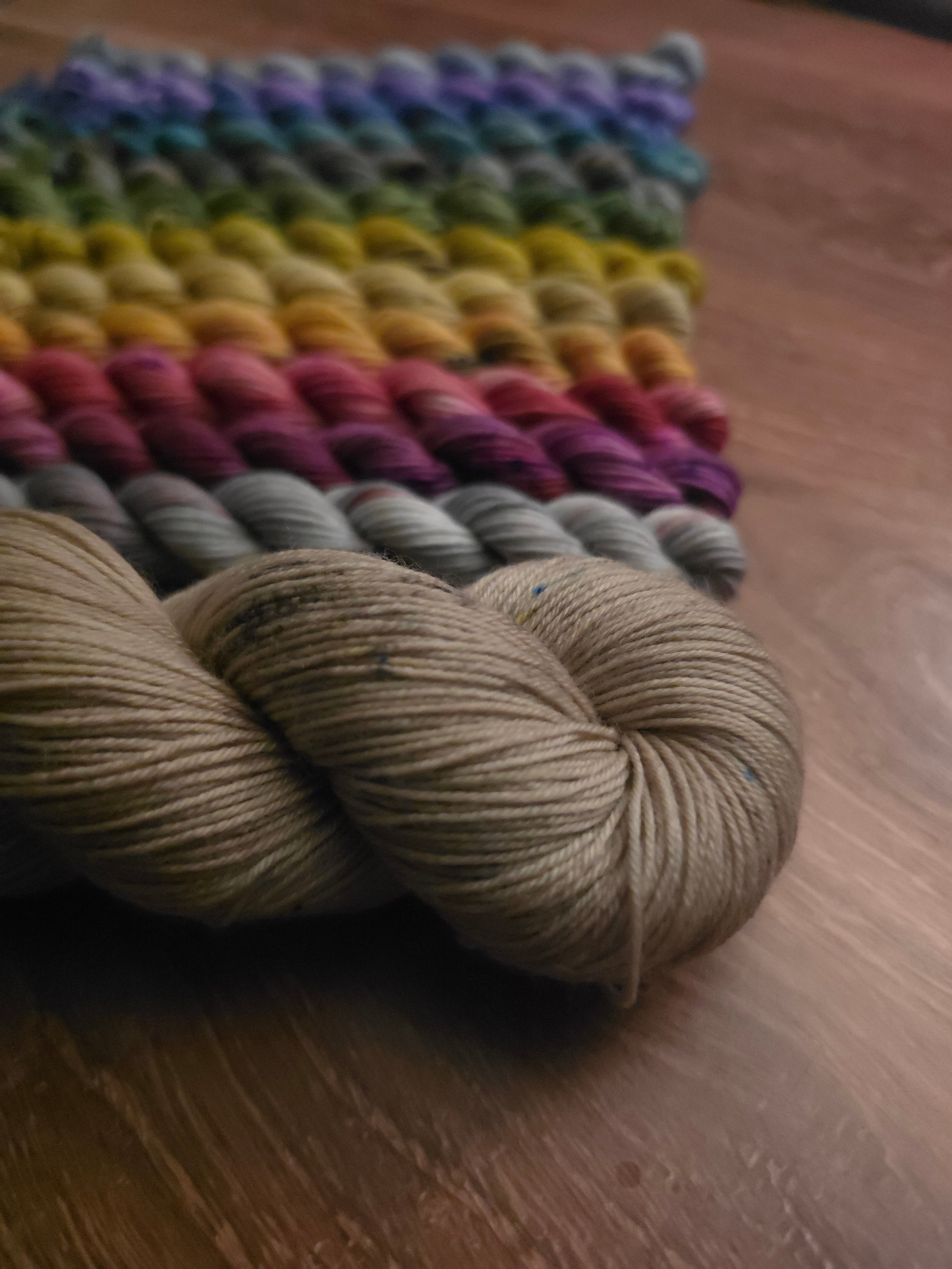 A close-up of a knitted textile made from variegated yarn in rainbow colors, with a skein of natural beige yarn in the foreground on a wooden surface.