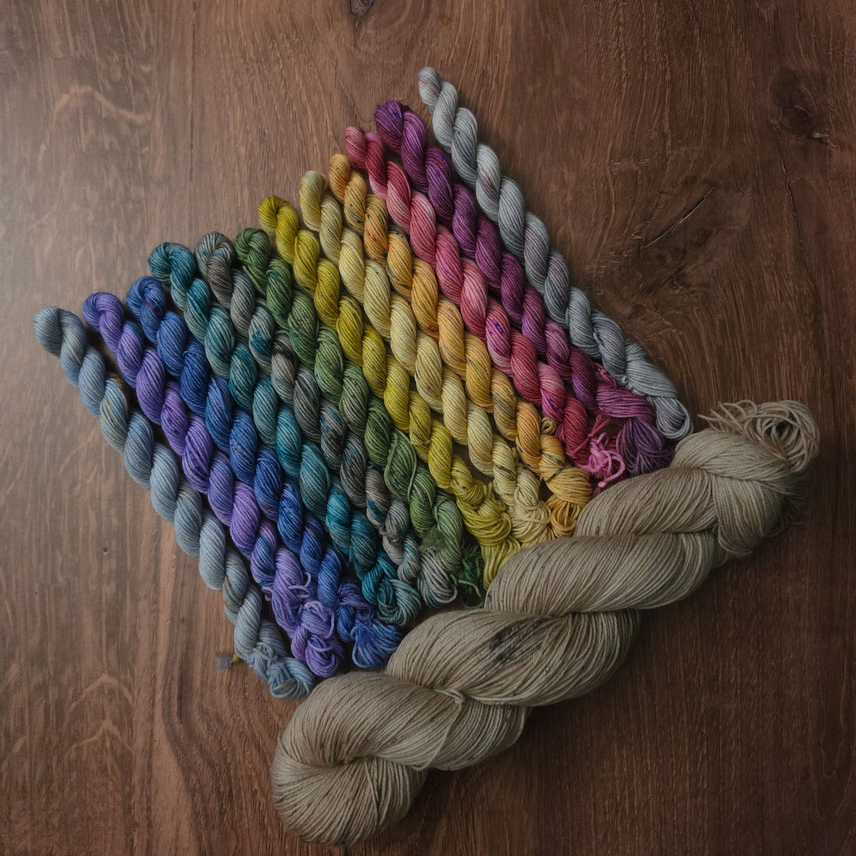 A set of twisted yarn skeins arranged in a fan shape on a dark wood background, showing a rainbow of colors from blue and purple through green, yellow, orange, pink, and lavender, with a large beige skein placed in front.